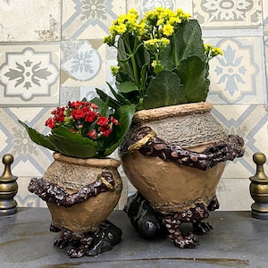 Könnte beinhalten: Zwei dekorative Keramiktöpfe, einer mit roten und der andere mit gelben Blumen. Die Töpfe haben ein strukturiertes, erdiges Design mit dunkelbraunen Akzenten und einem einzigartigen Sockel. Der Hintergrund zeigt eine gemusterte Fliesenwand.