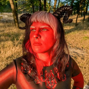 May include: A person with red face paint, black and white hair, and black horns. They are wearing a black top with a geometric design. The person has a nose ring and is looking off to the side.
