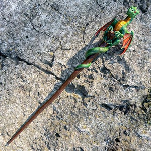 Puede incluir: Una varita mágica artesanal con un dragón verde y naranja. La varita es marrón con una vid verde enrollada. El dragón tiene alas rojas y escamas detalladas. La varita está sobre una superficie de piedra gris texturizada.
