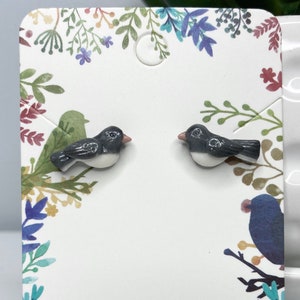 May include: A pair of gray and white ceramic bird earrings. The birds are facing to the right and have a small pink beak.