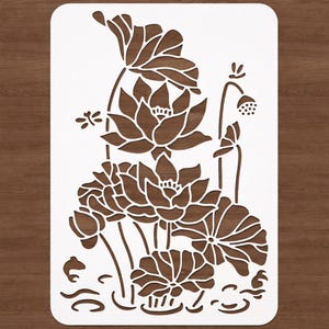 Puede incluir: Un esténcil blanco con un diseño de flor de loto marrón. El diseño incluye múltiples flores de loto, hojas y una vaina de semillas. El esténcil está sobre un fondo de madera.