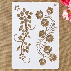 以下が含まれることがあります： 2つの花の模様が描かれた白いプラスチック製のステンシル。1つは花と葉の垂直配置です。もう1つは、湾曲した花のパターンです。「18」の数字が右上隅にあります。 クラフトやDIYプロジェクトに最適です。