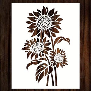 May include: A brown sunflower stencil design on a white background. The stencil features three sunflowers with detailed petals and centers, along with stems and leaves. The design is set against a dark wood grain background.