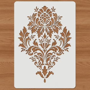 Könnte beinhalten: Eine weiße Schablone mit einem symmetrischen Damastmuster, das detaillierte Blumen- und Blattmuster aufweist. Die Schablone besteht aus einem langlebigen Material, das für verschiedene Bastelprojekte geeignet ist. Das Design ist rautenförmig.