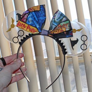 May include: A black headband with clear, round ears featuring gold wings and black glasses. A colorful bow with book covers and the words "Harry Potter" and "Fire" is centered on the headband. The ears have glitter.