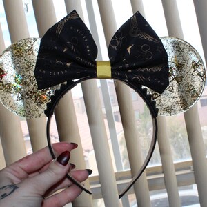 May include: A headband featuring two clear, circular ears filled with gold-toned, triangular, and circular metallic confetti. A black fabric bow with a gold-colored center knot sits between the ears, decorated with a repeating gold pattern of glasses, lightning bolts, and winged spheres.
