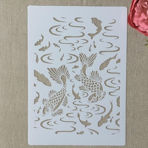 Könnte beinhalten: Weißer Kunststoff-Schablone mit einem Koi-Teich-Design. Die Schablone enthält detaillierte Ausschnitte von Koi-Fischen, Wasserwellen und kleineren Fischen. Geeignet für verschiedene Bastelprojekte.