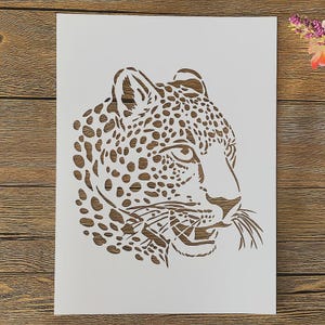 May include: A white stencil featuring a detailed profile of a leopard's head. The leopard's fur is represented by numerous brown spots. The stencil is on a wooden surface.