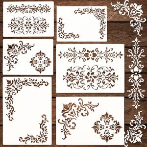 May include: A collection of white stencils with various ornate floral and scroll designs. The stencils include corner, border, and central motifs, perfect for decorative painting or crafting projects. The stencils are arranged on a wooden surface.