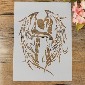 May include: A white stencil featuring a detailed design of two figures embracing, surrounded by large, stylized wings. The figures are in the center, with the wings extending outwards. The stencil is on a wooden surface.