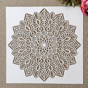 May include: A white mandala stencil with a detailed, symmetrical design. The stencil features a central floral pattern surrounded by layers of intricate, repeating shapes. The design is cut out, creating a negative space that can be used for painting or other crafts.