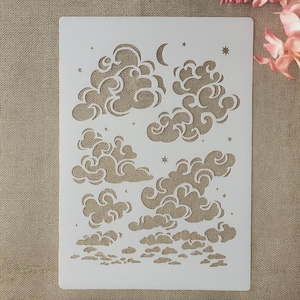 Pode incluir: Um estêncil de plástico branco com desenhos de nuvens e estrelas. O estêncil inclui várias formas de nuvens, uma lua crescente e várias estrelas pequenas. O desenho é recortado, permitindo que tinta ou outros meios sejam aplicados através das aberturas.
