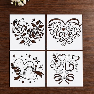 May include: Four white stencils with heart designs. One stencil features roses, another has the word "love" in cursive, a third has two hearts with a ribbon, and the last has heart-shaped balloons.