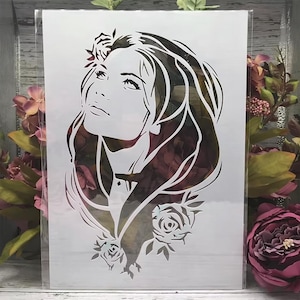 Puede incluir: Un esténcil blanco que presenta el perfil de una mujer con cabello largo y detalles florales. El diseño incluye rosas en la parte inferior y una corona floral. El fondo muestra una mezcla de colores burdeos y verdes.