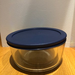 May include: A clear glass storage container with a blue lid. The container is empty and sitting on a wooden surface.