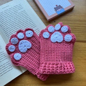 May include: A pair of pink crocheted fingerless gloves with white paw print designs. The gloves are made with a soft, textured yarn.