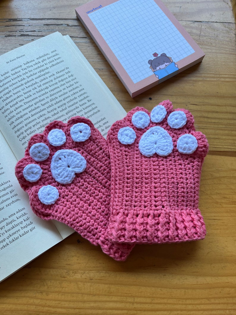 May include: A pair of pink crocheted fingerless gloves with white paw print designs. The gloves are made with a soft, textured yarn.