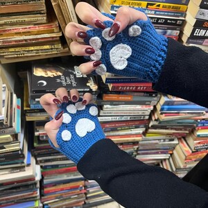 Pu&ograve; includere: Un paio di guanti senza dita blu con impronte di zampe bianche. I guanti sono all'uncinetto e hanno un polsino nero.