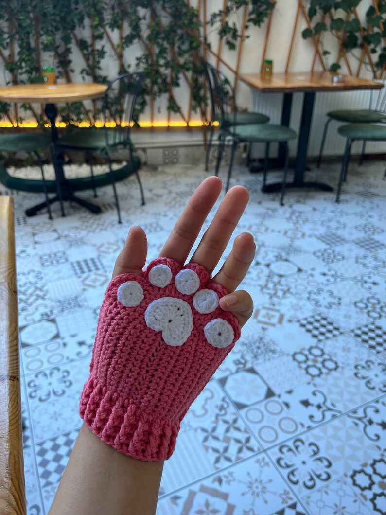 May include: Pink crocheted fingerless gloves with a white paw print design.