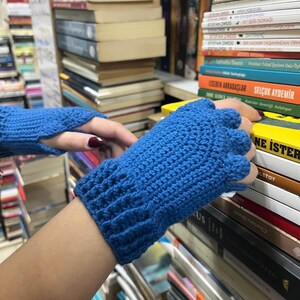Pu&ograve; includere: Un paio di guanti senza dita blu realizzati in filato all'uncinetto. I guanti sono mostrati su uno sfondo di librerie piene di libri.
