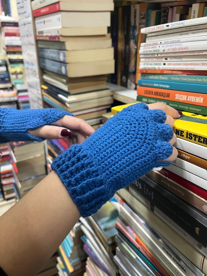 Pu&ograve; includere: Un paio di guanti senza dita blu realizzati in filato all'uncinetto. I guanti sono mostrati su uno sfondo di librerie piene di libri.