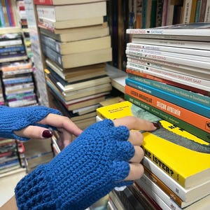Pu&ograve; includere: Un paio di guanti senza dita blu fatti a crochet. I guanti sono sopra una pila di libri con il titolo "NE ISTER?" visibile.