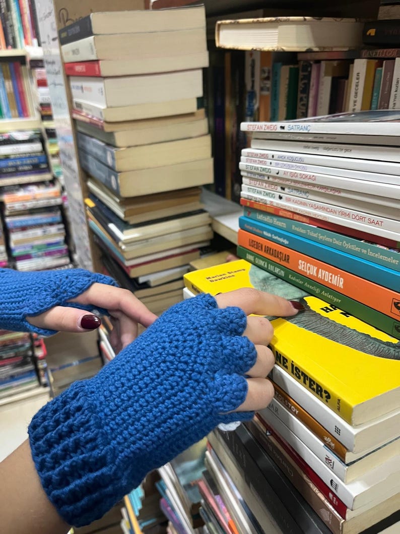 Pu&ograve; includere: Un paio di guanti senza dita blu fatti a crochet. I guanti sono sopra una pila di libri con il titolo "NE ISTER?" visibile.