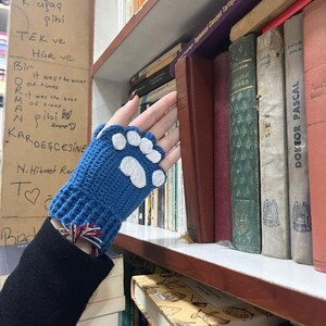 Pu&ograve; includere: Guanti senza dita in crochet blu con disegni di impronte di zampe bianche. I guanti sono su una mano che raggiunge una libreria piena di libri.