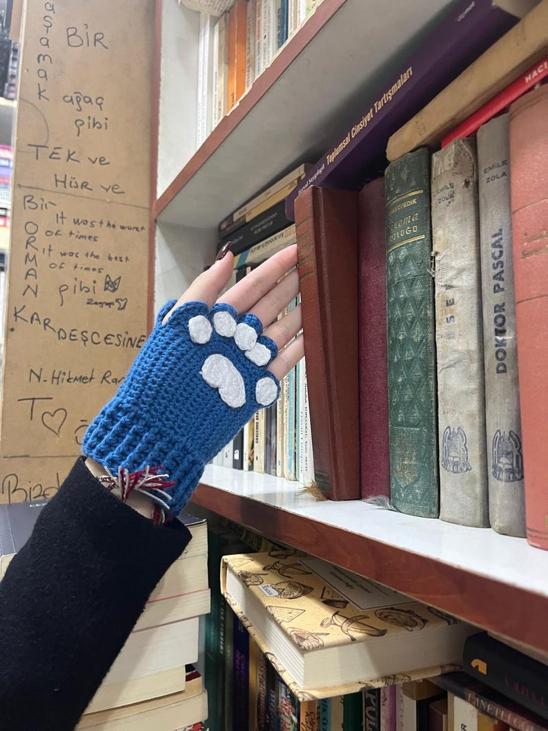 Pu&ograve; includere: Guanti senza dita in crochet blu con disegni di impronte di zampe bianche. I guanti sono su una mano che raggiunge una libreria piena di libri.