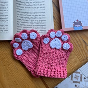 May include: A pair of pink crocheted fingerless gloves with white paw print accents. The gloves are on a wooden surface.