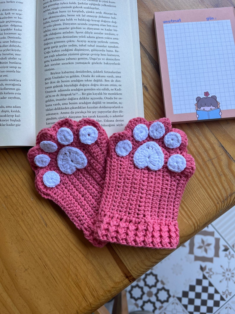 May include: A pair of pink crocheted fingerless gloves with white paw print accents. The gloves are on a wooden surface.