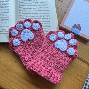 May include: A pair of pink crocheted mittens with white paw print designs. The mittens are made with a soft, textured yarn and have a comfortable fit.