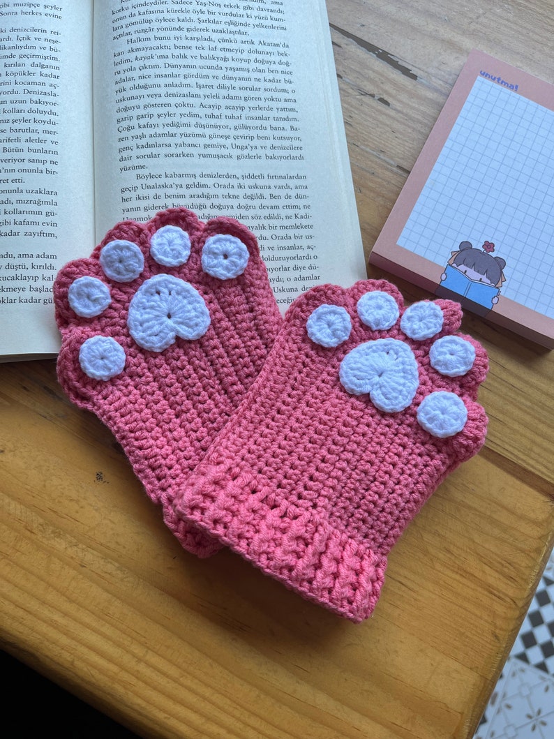 May include: A pair of pink crocheted mittens with white paw print designs. The mittens are made with a soft, textured yarn and have a comfortable fit.