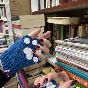 Pu&ograve; includere: Un paio di guanti senza dita di lana blu con disegni di impronte di zampa bianche. I guanti sono mostrati contro una libreria con vari libri.