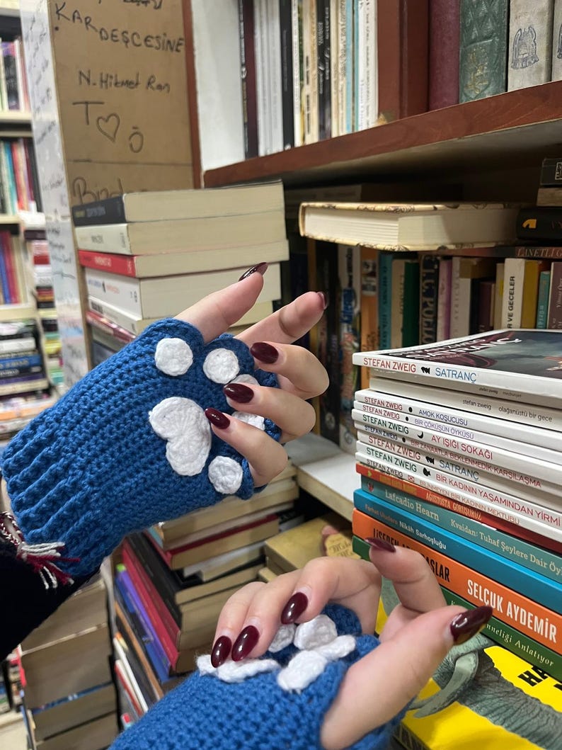 Pu&ograve; includere: Un paio di guanti senza dita di lana blu con disegni di impronte di zampa bianche. I guanti sono mostrati contro una libreria con vari libri.