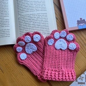 May include: A pair of pink crocheted fingerless gloves with white paw print designs. The gloves are lying on a wooden surface.