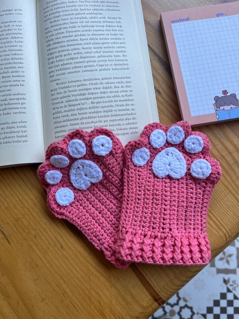 May include: A pair of pink crocheted fingerless gloves with white paw print designs. The gloves are lying on a wooden surface.