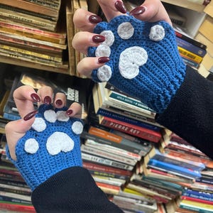 Pu&ograve; includere: Un paio di guanti senza dita di colore blu all'uncinetto con disegni di impronte di zampe bianche. I guanti sono indossati da una persona con smalto scuro.