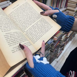 Pu&ograve; includere: Primo piano delle mani di una persona che indossa guanti senza dita blu e bianchi. La persona tiene un libro aperto a una pagina con testo turco. Il libro &egrave; su uno scaffale di libri.