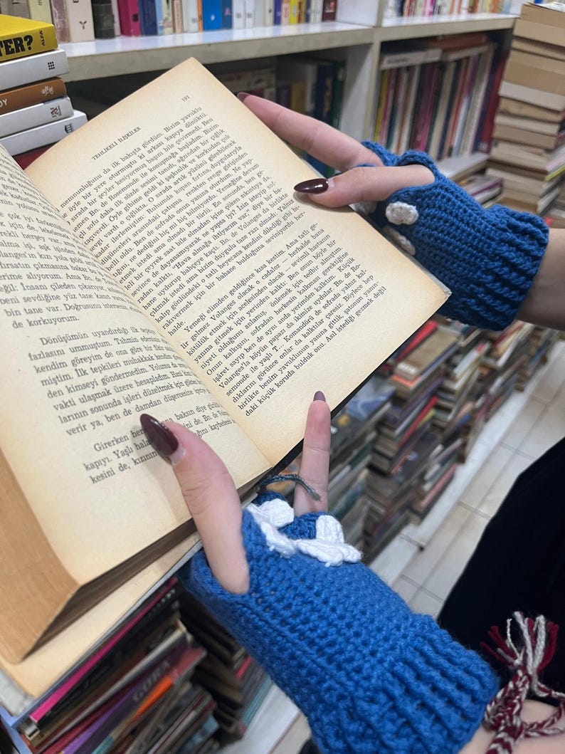 Pu&ograve; includere: Primo piano delle mani di una persona che indossa guanti senza dita blu e bianchi. La persona tiene un libro aperto a una pagina con testo turco. Il libro &egrave; su uno scaffale di libri.