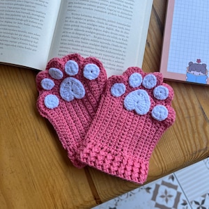 May include: A pair of pink crocheted mittens with white paw print details. The mittens are on a wooden surface.