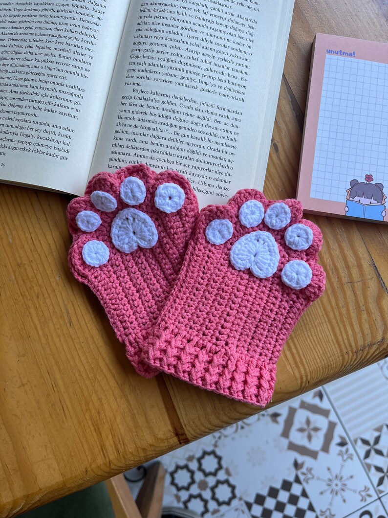 May include: A pair of pink crocheted mittens with white paw print details. The mittens are on a wooden surface.
