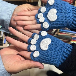 Pu&ograve; includere: Un paio di guanti senza dita blu con disegni di impronte di zampe bianche. I guanti sono all'uncinetto e hanno un aspetto testurizzato.