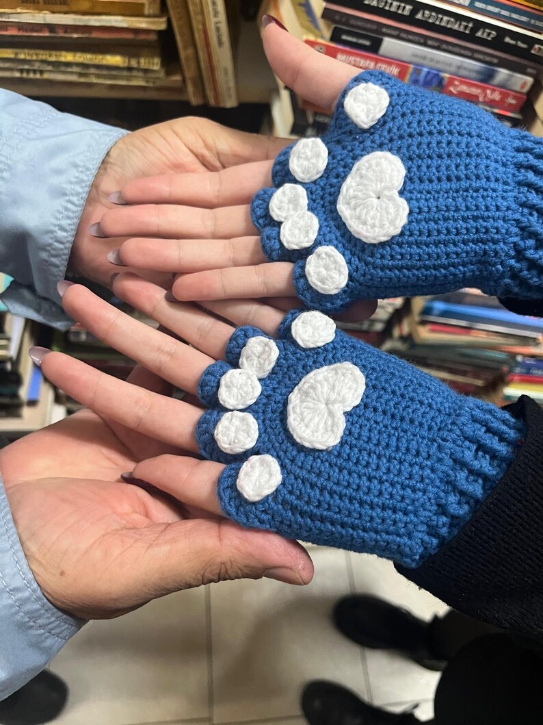 Pu&ograve; includere: Un paio di guanti senza dita blu con disegni di impronte di zampe bianche. I guanti sono all'uncinetto e hanno un aspetto testurizzato.