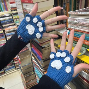 Pu&ograve; includere: Un paio di guanti senza dita blu con disegni di impronte di zampa bianche. I guanti sono all'uncinetto e hanno un aspetto testurizzato.