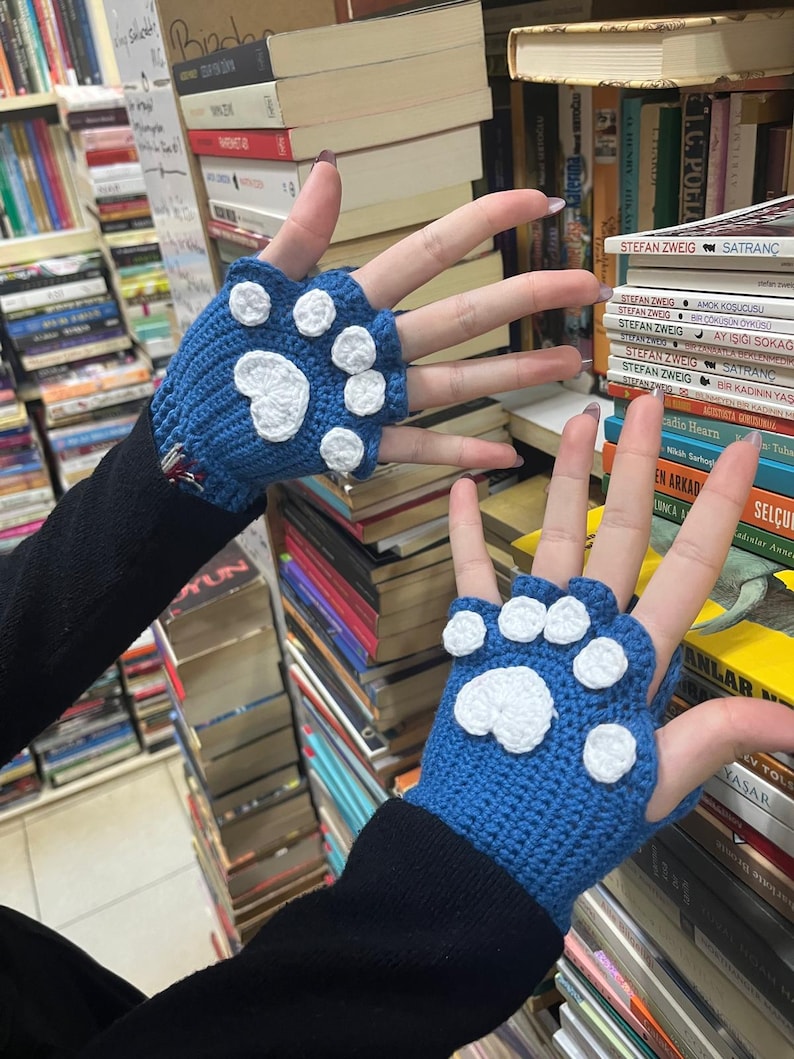Pu&ograve; includere: Un paio di guanti senza dita blu con disegni di impronte di zampa bianche. I guanti sono all'uncinetto e hanno un aspetto testurizzato.