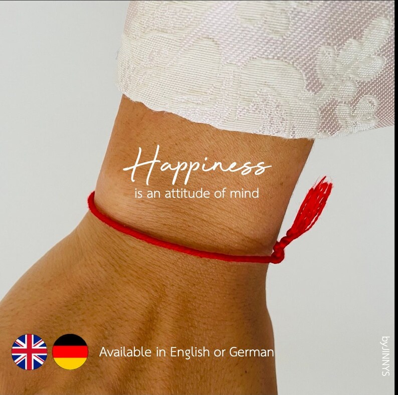 May include: Red string bracelet with a tassel on a person's wrist. The bracelet has the text "Happiness is an attitude of mind" written on it. The bracelet is available in English or German.
