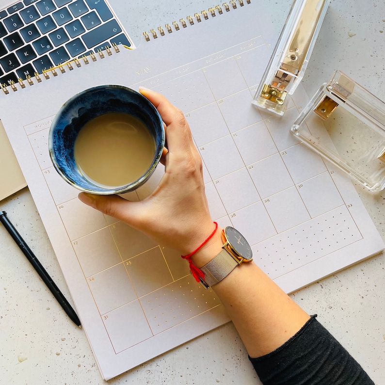 May include: A hand holding a blue and white ceramic mug filled with a light brown beverage. A red string bracelet is worn on the wrist of the hand. A gold watch with a black face is on the wrist. A white calendar with gold numbers is in the background.