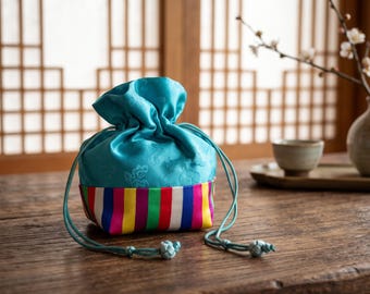 May include: A turquoise drawstring pouch featuring a subtle floral pattern on the upper section and a base decorated with vertical stripes in vibrant shades of pink, yellow, green, purple, and white, finished with matching turquoise cords.