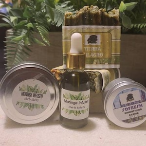 May include: Three small jars of body care products with green and white labels. The labels read "Moringa Infused Body Butter", "Moringa Infused Hair & Body Oil", and "Sofresh Smoothing Creme". A bar of soap with a green and gold label that reads "Tierra Milagro" is in the background.